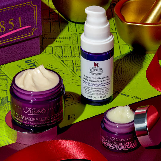 Drie Kiehl's Super Multi-Corrective producten – een cremepot, een oogcreme en een serumpotje – zijn gerangschikt op een rood lint, naast een paarse doos en gouden feestelijke decoraties, tegen een limoengroene achtergrond met een kaartpatroon.