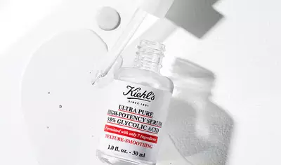 A clear bottle of Kiehl's Ultra Pure High-Potency Serum with 38% Glycolic Acid, partially spilled, on a light background. The bottle is labeled with product information, emphasizing texture-smoothing benefits.