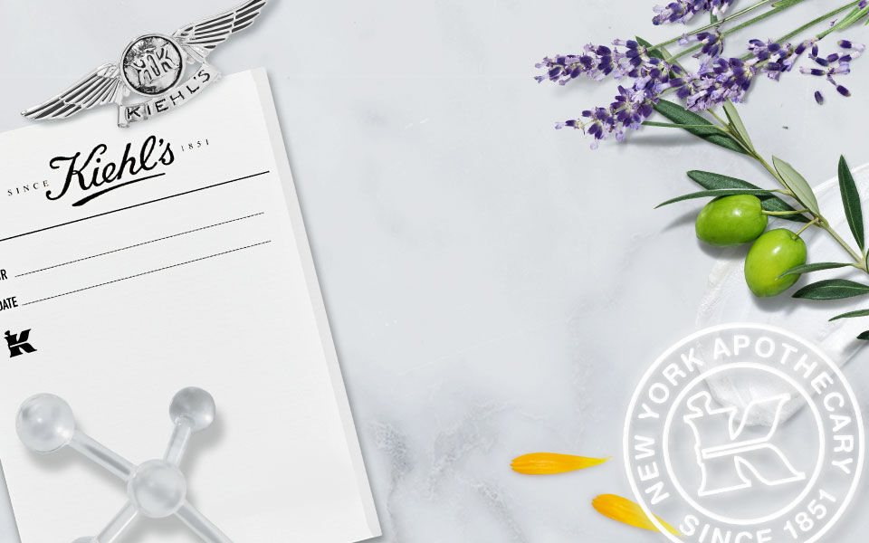 Een collage met een historische foto van de Kiehl's apotheek, omgeven door lavendel, olijven en bloemblaadjes, met het Kiehl's logo en de tekst 'New York Apothecary Since 1851'.