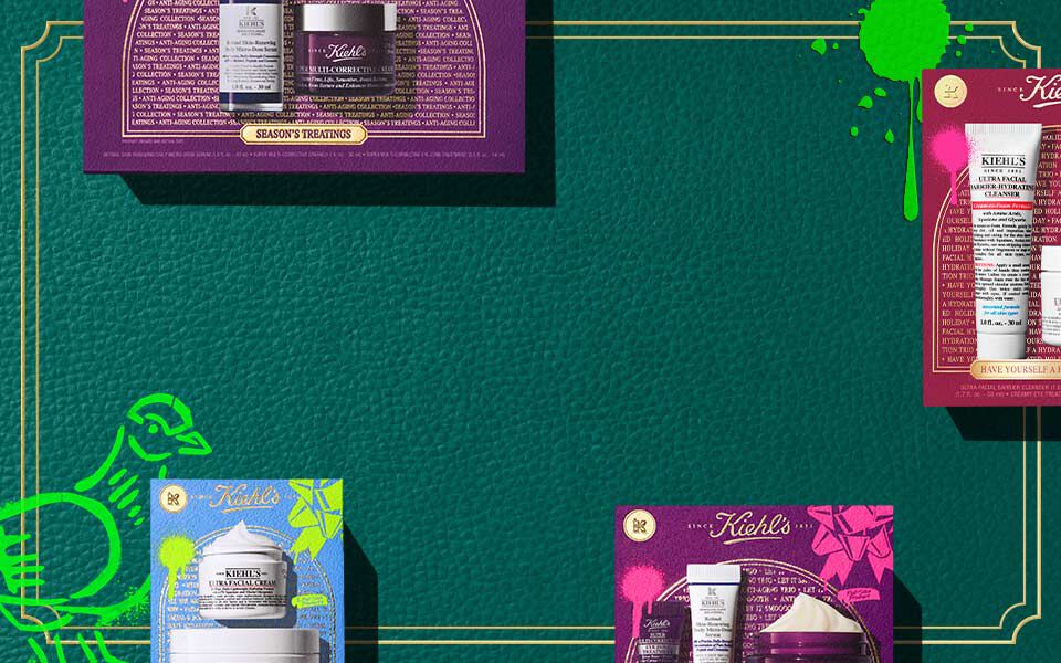 Horizontale banner met een donkergroene getextureerde achtergrond en gouden randlijnen, met kleurrijke Kiehl's geschenkdozen in roze, blauw en paars, die elk huidverzorgingsproducten tonen. Groene artistieke accenten verschijnen aan beide zijden.