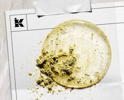 Een cirkelvormige veeg lichte creme met een gestructureerd hoopje groen poeder, gepresenteerd op een wit vel met een logo.