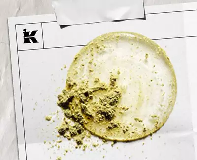Een cirkelvormige veeg lichte creme met een gestructureerd hoopje groen poeder, gepresenteerd op een wit vel met een logo.