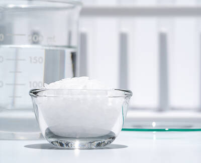 A small glass bowl filled with white granules sits on a table next to a measuring beaker containing clear liquid. In the background, a white surface provides a simple backdrop.