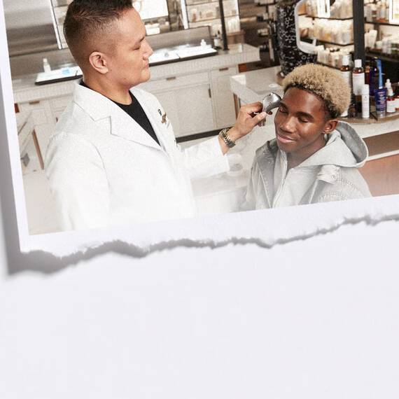 Een man in een laboratoriumjas gebruikt een zilverkleurig handapparaat om een huidbehandeling uit te voeren op een lachende jongeman met licht krullend haar, wiens ogen gesloten zijn. De scene bevindt zich in wat lijkt op een winkel- of laboratoriumomgeving met productrekken op de achtergrond, en het beeld is omlijst met een gescheurd papiereffect aan de onderkant.