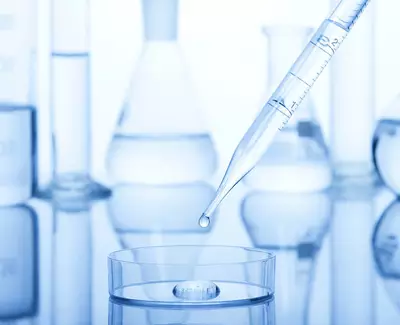 A dropper releasing a drop of liquid into a petri dish, surrounded by laboratory glassware including beakers and flasks, all set against a blue-tinted background.