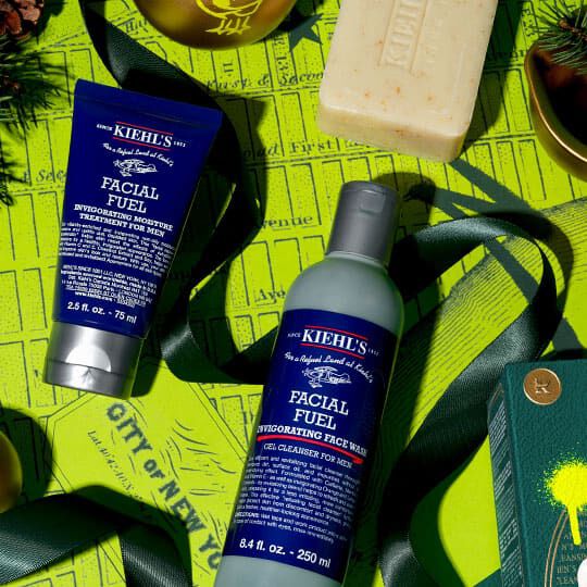 Kiehl's Facial Fuel reiniger en moisturizer gerangschikt met een stuk zeep en groene kerstdecoraties op een felgele achtergrond.
