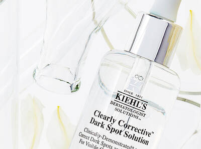 Fles met Kiehl's Clearly Corrective Dark Spot Solution op een lichte achtergrond met bloemen en glaswerk.