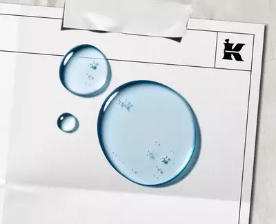 Twee druppels water op een leeg vel papier met een zwart "K"-logo en een stuk tape bovenaan.