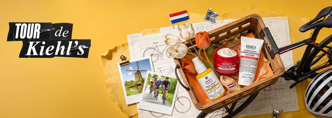 Drie Kiehl's huidverzorgingsproducten opgesteld tegen een felgele achtergrond met een wielrenthema. Links staat een gele pompfles van het "Better Screen UV Serum", in het midden een geopende rode pot met de "Ultra Facial Meltdown Recovery Cream" die een rijke witte creme laat zien, en rechts een witte tube van de "Ultra Facial Barrier-Hydrating Cleanser". Op de achtergrond liggen een witte fietshelm met de merknaam "Kiehl's" en een sportieve zonnebril. Rondom de producten liggen een landkaart, decoratieve miniatuurfietsen en zwart-wit polaroidfoto's van fietsers.