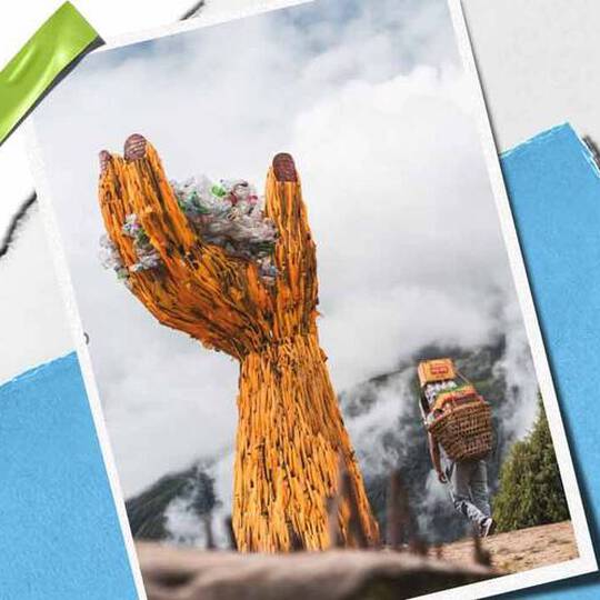 Een torenhoge, houten handsculptuur houdt een stapel afval vast, als symbool voor milieubewustzijn, met in de buurt een persoon die goederen draagt.