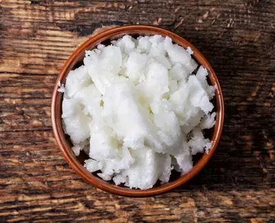 A wooden bowl filled with fluffy white coconut or shea butter, placed on a textured wooden surface.