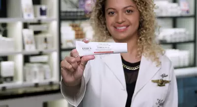A person with curly hair holds a tube of Kiehl's Barrier Cream, smiling in a store filled with skincare products.