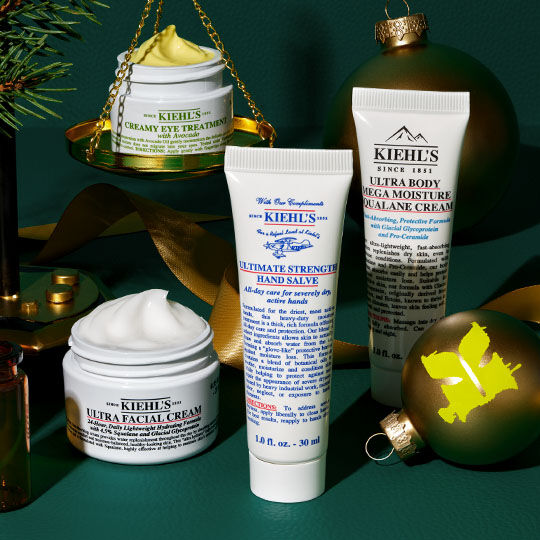 Een feestelijke opstelling van Kiehl's producten, waaronder Creamy Eye Treatment op gouden weegschalen, Ultra Facial Cream, Ultimate Strength Hand Salve en Ultra Body Mega Moisture Squalane Cream, omringd door groene ornamenten, dennentakken en gouden linten op een donkergroene achtergrond.