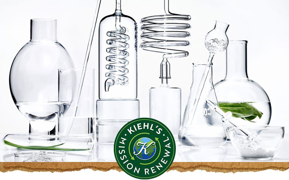 Een verzameling doorzichtige glazen laboratoriumvaten gevuld met water en een groen blad, met het logo van Kiehl's aan de rechterkant.