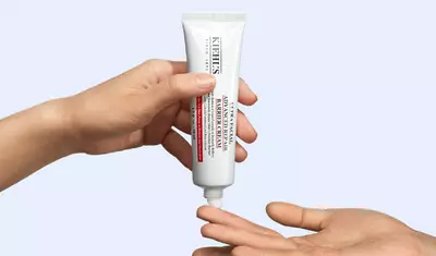 A hand is holding a tube of Kiehl's skincare cream, while another hand is positioned below to receive the product. The background is a soft blue.