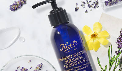 A blue bottle of Kiehl's Midnight Recovery Botanical Cleansing Oil with a pump top, surrounded by lavender flowers, a yellow flower, and droplets of oil on a marble surface.