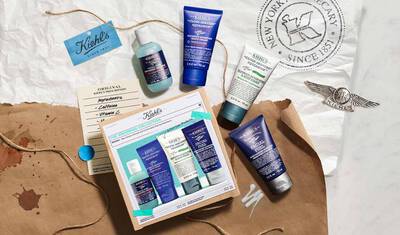 A flat lay of Kiehl's skincare products arranged aesthetically on a textured surface, featuring various tubes and bottles alongside branded packaging and a label with ingredients.