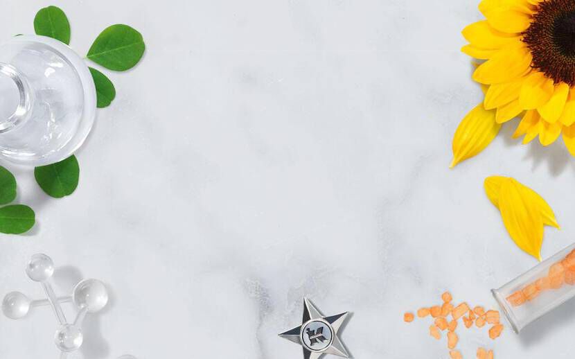 A flat lay of various natural elements, including sunflower petals, green olives, leaves, and floral elements, arranged around laboratory glassware and a star-shaped decoration on a marble surface.