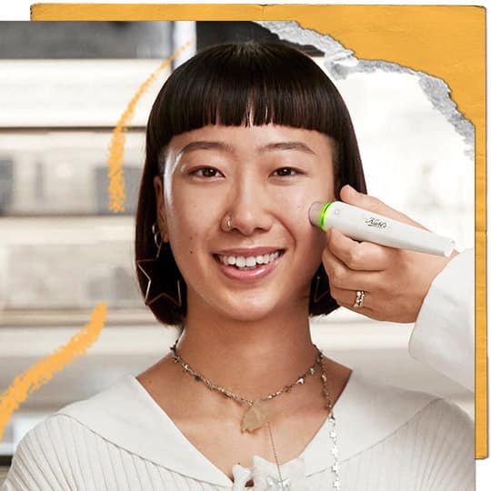 Een lachend persoon met kort donker haar en ster-oorbellen heeft een wit Kiehl's-apparaat met een gloeiende groene punt aangebracht op zijn wang door een hand. De achtergrond bevat planken en de afbeelding is omlijst met gescheurd geel papier en oranje lijnen.