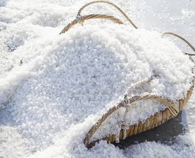 A woven basket filled with white salt sits on a beach, with salt crystals scattered around it glistening in the sunlight.