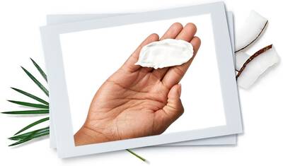 A close-up of a hand holding a small amount of cream, set against a white background with coconut pieces and green leaves nearby.