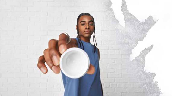 Een persoon met een blauw mouwloos shirt houdt een kleine witte kom in de richting van de camera, tegen een witte muur met textuur.