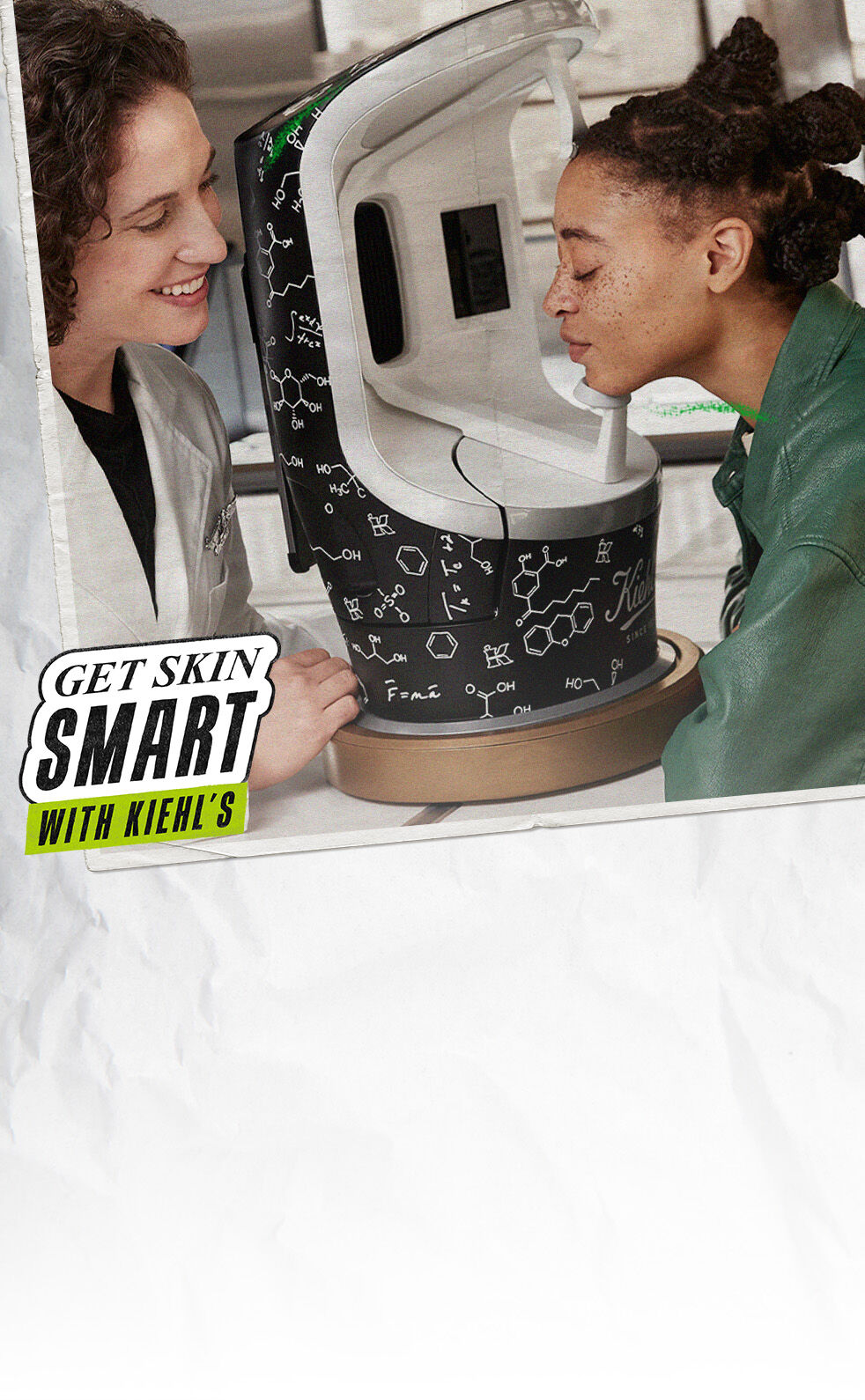 Een vrouw en een jongere persoon staan naast een huidanalyse-apparaat van Kiehl's. De vrouw legt iets uit terwijl de jongere persoon het apparaat beziet.