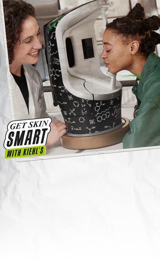 Een vrouw en een jongere persoon staan naast een huidanalyse-apparaat van Kiehl's. De vrouw legt iets uit terwijl de jongere persoon het apparaat beziet.