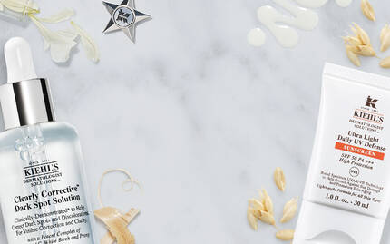 Alt-text in Dutch: "Twee Kiehl's huidverzorgingsproducten op een marmeren ondergrond, inclusief een serum en een creme, omgeven door natuurlijke ingredienten zoals haver."