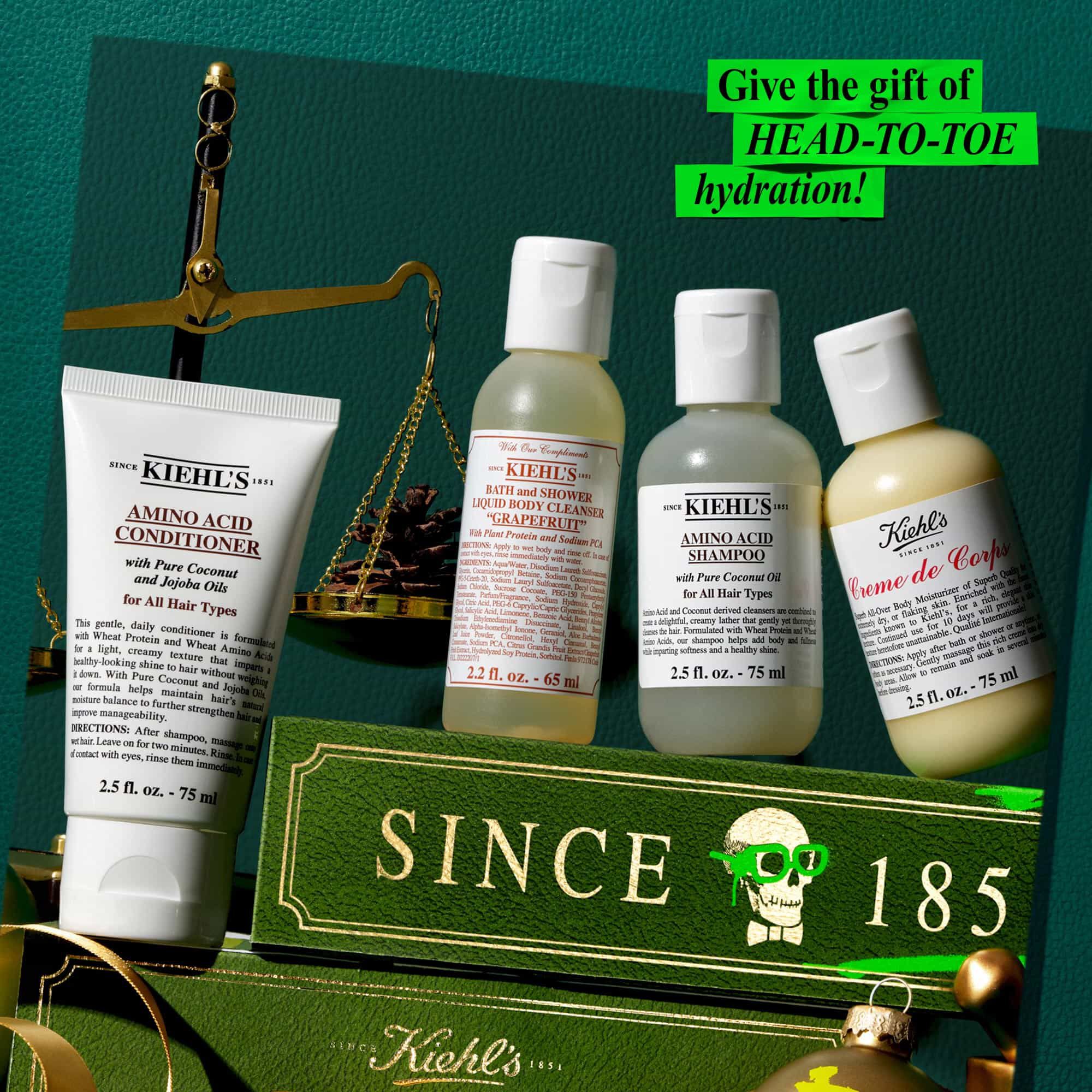 Tegen een donkergroene achtergrond met gouden en groene accenten staan vier producten rondom een groene en gouden geschenkverpakking met de tekst "SINCE 1851". De producten bestaan uit een tube "KIEHL'S AMINO ACID CONDITIONER", een fles "KIEHL'S BATH AND SHOWER LIQUID BODY CLEANSER GRAPEFRUIT", een fles "KIEHL'S AMINO ACID SHAMPOO" en een fles "KIEHL'S CREME DE CORPS". Linksboven zijn een gouden balans en een kleine dennenappel te zien. In een felgroen vakje in de rechterbovenhoek staat de tekst "Geef het cadeau van hydratatie van HEAD-TO-TOE!".