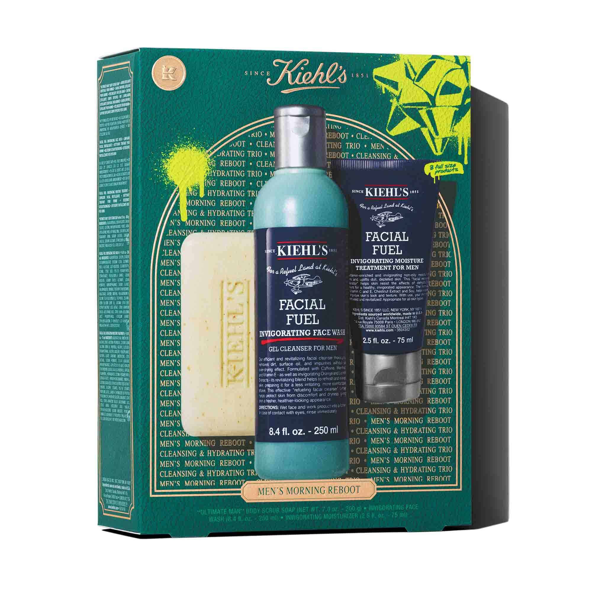 Een afbeelding van een donker groenblauwe geschenkdoos van Kiehl's met witte en gouden accenten. De doos bevat drie producten: een reep "KIEHL'S 'ULTIMATE MAN' BODY SCRUB SOAP", een blauwe fles "KIEHL'S FACIAL FUEL ENERGIZING FACE WASH" en een donkerblauwe tube "KIEHL'S FACIAL FUEL MOISTURIZER". De doos is versierd met felgroene grafische verfspatten en een strikontwerp en heeft onderaan de tekst "MEN'S MORNING REBOOT".