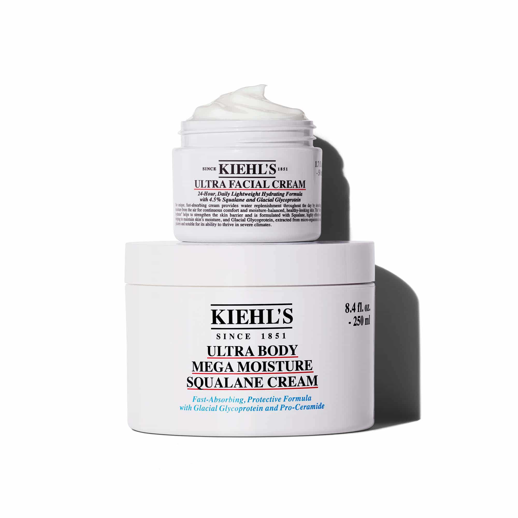 Een afbeelding met twee witte potten Kiehl's creme, op elkaar gestapeld, die een schaduw werpen tegen een witte achtergrond. De kleinere bovenste pot is "KIEHL'S ULTRA FACIAL CREAM" met een zichtbare werveling van witte creme. De grotere onderste pot is "KIEHL'S ULTRA BODY MEGA MOISTURE SQUALANE CREAM".