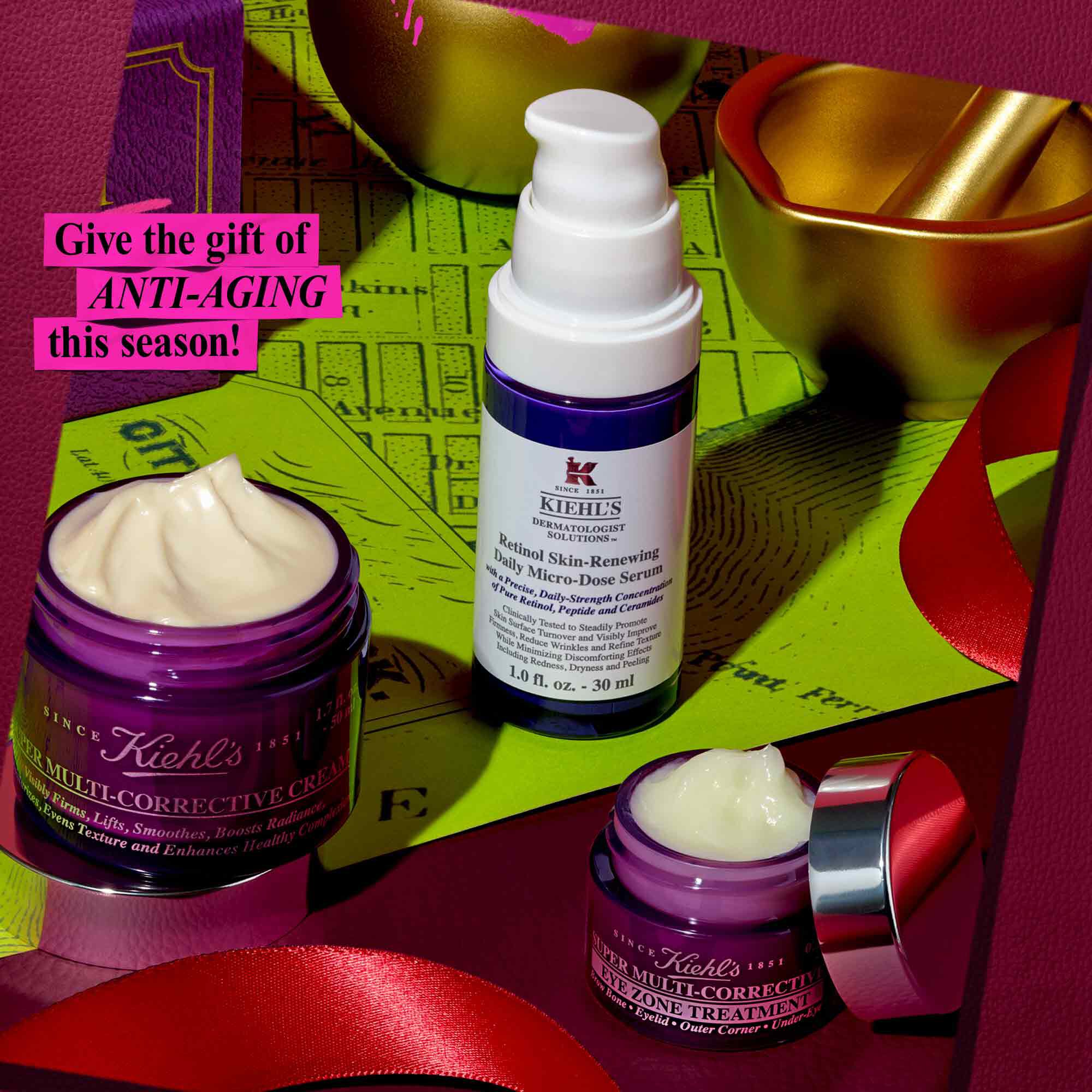 Een feestelijke afbeelding toont drie anti-verouderingsproducten van Kiehl's: een paarse pot met "Kiehl's Super Multi-Corrective Cream", een witte en paarse fles met "Kiehl's Retinol Skin-Renewing Daily Micro-Dose Serum" en een kleinere paarse pot met "Kiehl's Super Multi-Corrective Eye Zone Treatment". Ze zijn gerangschikt op een groene ondergrond, met rode linten en een gouden vijzel en stamper op de achtergrond. In een roze tekstkader in de linkerbovenhoek staat: "Geef het cadeau van ANTI-AGING dit seizoen!".