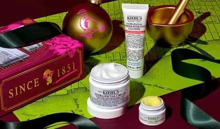 Een feestelijke afbeelding van een witte tube "KIEHL'S ULTRA FACIAL BARRIER-HYDRATING CLEANSER", een witte pot "KIEHL'S ULTRA FACIAL CREAM" met zichtbare witte creme en een kleine witte pot "KIEHL'S CREAMY EYE TREATMENT WITH AVOCADO" met zichtbare lichtgele creme, allemaal gerangschikt op een donkerrode en groene achtergrond met een landkaartpatroon. Rechtsboven is een gouden vijzel en stamper zichtbaar. Linksboven zijn een gouden kerstornament met een roze vogelontwerp en een rode geschenkdoos met "SINCE 1851" en een doodshoofdontwerp aanwezig. Rond de producten is zwart lint gedrapeerd.