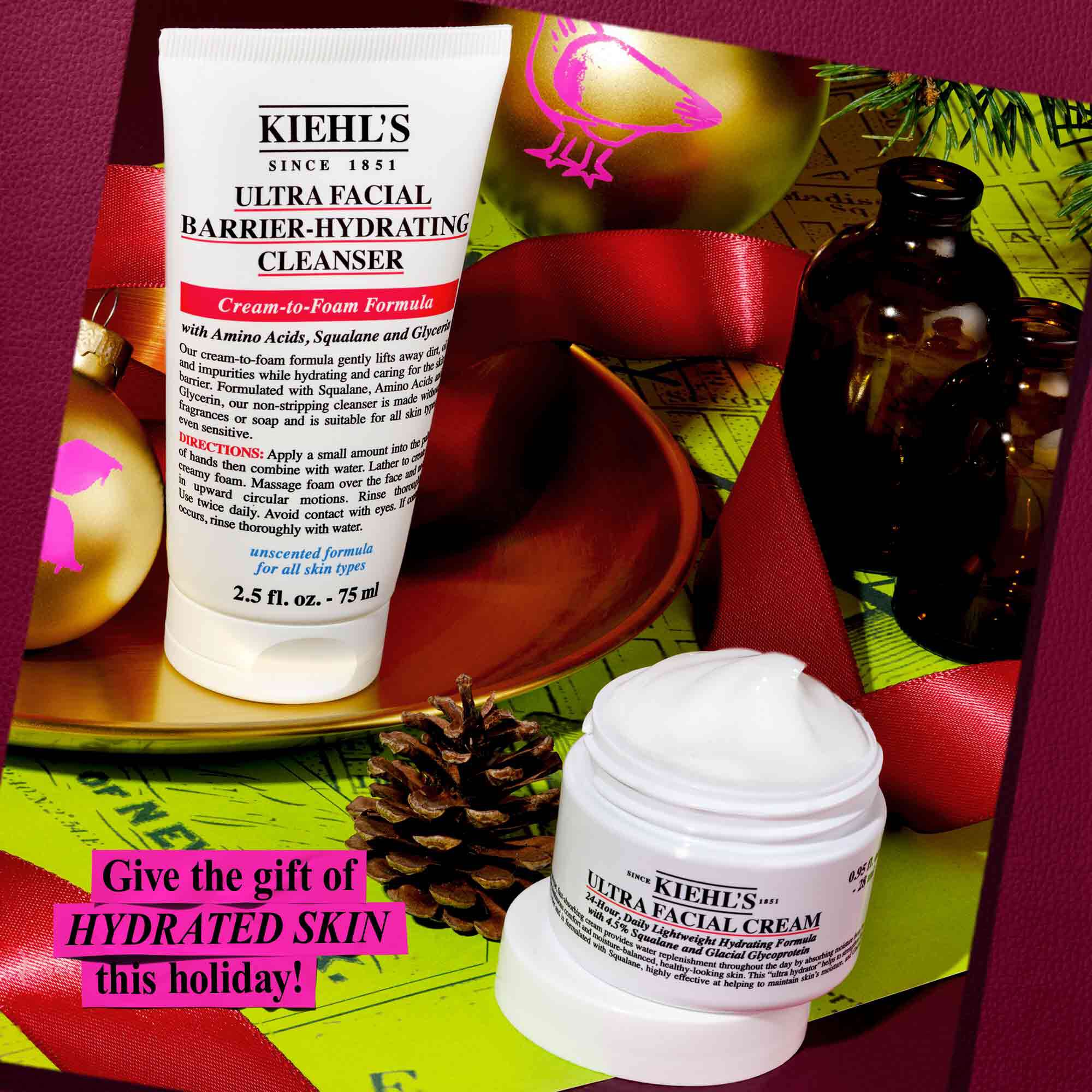 Een feestelijke afbeelding van een witte tube Kiehl's Ultra Facial Barrier-Hydrating Cleanser en een witte pot Kiehl's Ultra Facial Cream, tegen een achtergrond van rood lint en groen inpakpapier met gouden ornamenten. Op een prominente plaats staat de tekst: "Geef deze feestdagen een gehydrateerde huid cadeau!