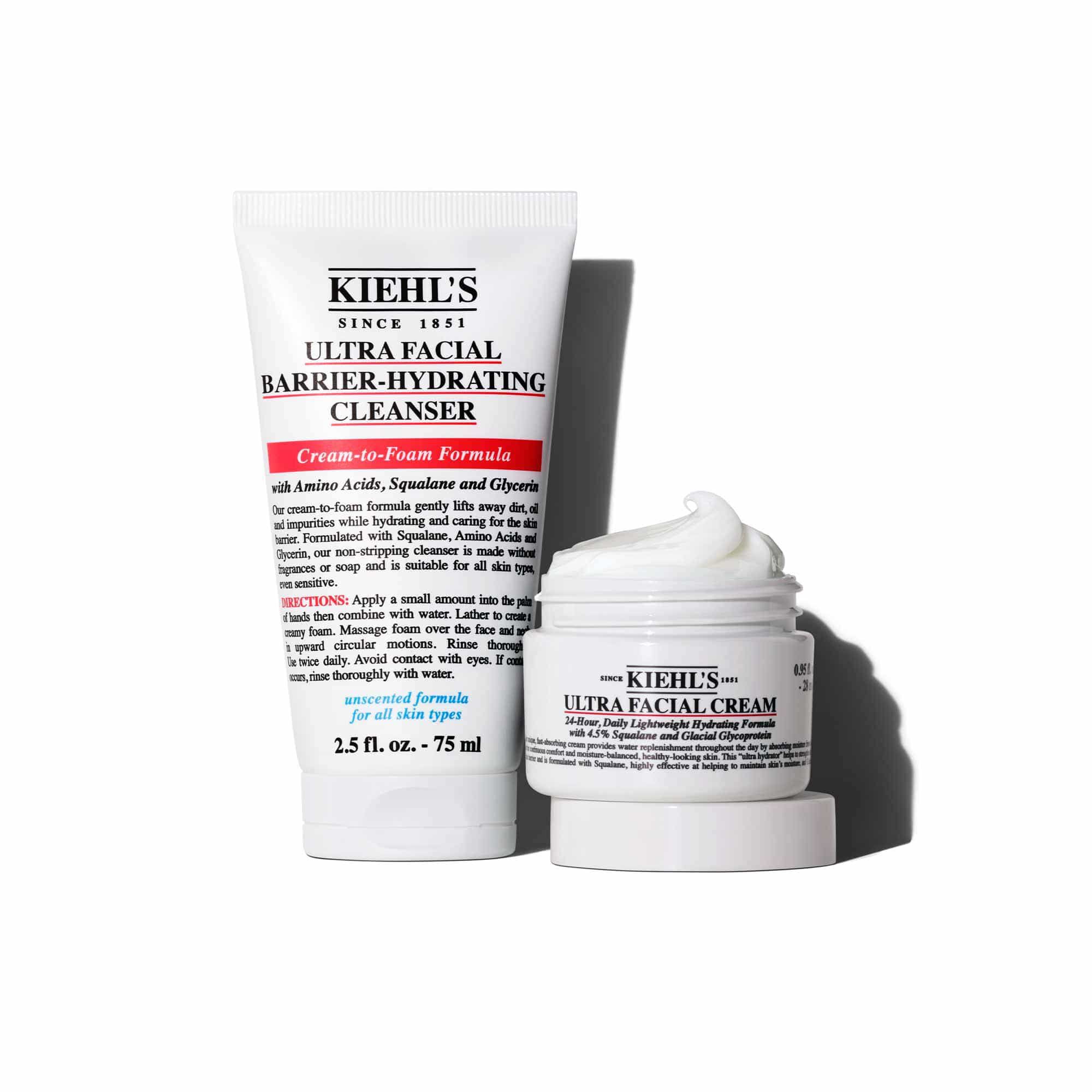 Een afbeelding toont twee Kiehl's producten: een witte tube "KIEHL'S ULTRA FACIAL BARRIER-HYDRATING CLEANSER", rechtopstaand naast een geopende witte pot "KIEHL'S ULTRA FACIAL CREAM". Het deksel van de pot is eronder geplaatst en de hele set staat op een witte achtergrond.