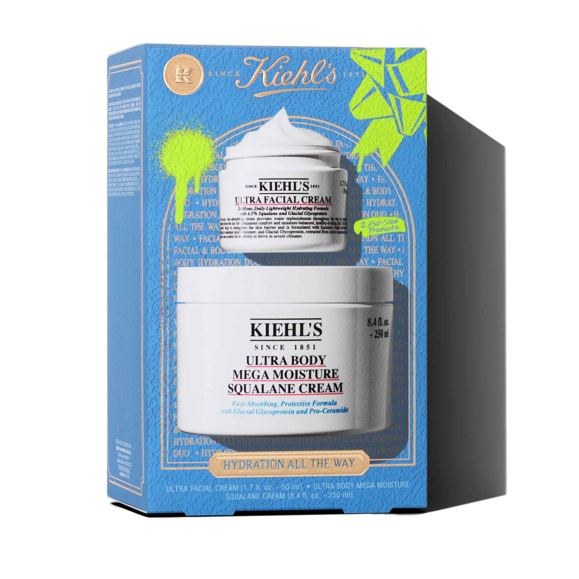 Een afbeelding van een blauwe geschenkdoos van Kiehl's. De doos bevat twee witte potten: "KIEHL'S ULTRA FACIAL CREAM" en "KIEHL'S ULTRA BODY MEGA MOISTURE SQUALANE CREAM." De doos heeft felgroene grafische verfspatten en een strikontwerp, en op de voorkant staat de tekst "HYDRATION ALL THE WAY".