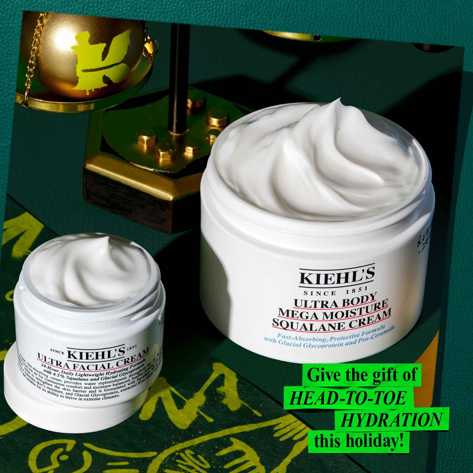 Een afbeelding tegen een donkergroene achtergrond met gouden en groene accenten toont twee geopende witte potten met zichtbare productdruppels. De kleinere pot is "KIEHL'S ULTRA FACIAL CREAM" en de grotere pot is "KIEHL'S ULTRA BODY MEGA MOISTURE SQUALANE CREAM". Linksboven staat een gouden kerstversiering met een groen "K"-logo. In de rechterbenedenhoek staat in heldergroene tekst "Geef deze feestdagen HEAD-TO-TOE HYDRATION cadeau!".