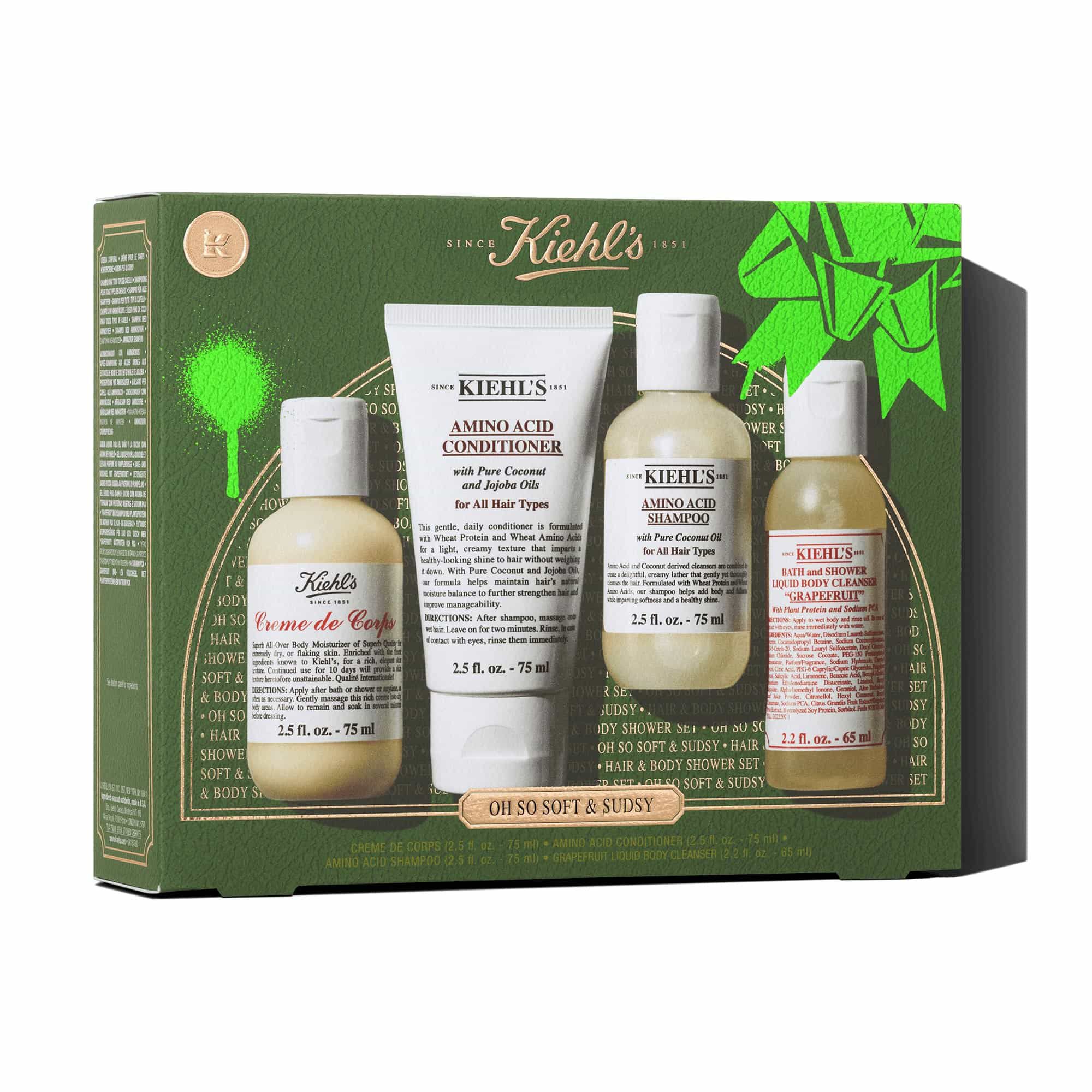 Een afbeelding tegen een donkergroene achtergrond met structuur toont vier producten. De bovenste rij toont: een fles "KIEHL'S CREME DE CORPS", een tube "KIEHL'S AMINO ACID CONDITIONER", een fles "KIEHL'S AMINO ACID SHAMPOO" en een fles "KIEHL'S BATH AND SHOWER LIQUID BODY CLEANSER GRAPEFRUIT". Daaronder toont een groene Kiehl's geschenkverpakking dezelfde vier producten netjes gerangschikt binnenin. De geschenkverpakking heeft een heldergroen grafisch accent.