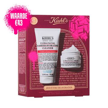 Een afbeelding toont een doos met een geschenkset van Kiehl's, voornamelijk in donkerrood, met rechtsboven een roze strik en linksonder een roze verfspetter. Op de voorkant van de doos staat "DUO IT FOR THE HYDRATION", samen met "ULTRA FACIAL BARRIER-HYDRATING CLENSER + ULTRA FACIAL CREAM". Rechts van de doos zijn de twee producten buiten hun verpakking afgebeeld.