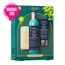 Een afbeelding van een donker groenblauwe geschenkdoos van Kiehl's met witte en gouden accenten aan de linkerkant. De doos bevat drie producten. Rechts van de doos staan dezelfde drie producten buiten de doos: een reep "KIEHL'S 'ULTIMATE MAN' BODY SCRUB SOAP", een blauwe fles "KIEHL'S FACIAL FUEL ENERGIZING FACE WASH" en een donkerblauwe tube "KIEHL'S FACIAL FUEL MOISTURIZER". De doos is versierd met felgroene grafische verfspatten en een strikontwerp en bevat de tekst "MEN'S MORNING REBOOT" aan de onderkant.